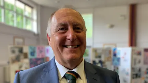 Adrian Whiteley is wearing a grey suit and green, gold, black and red striped tie. He is standing in front of a classroom art display. 